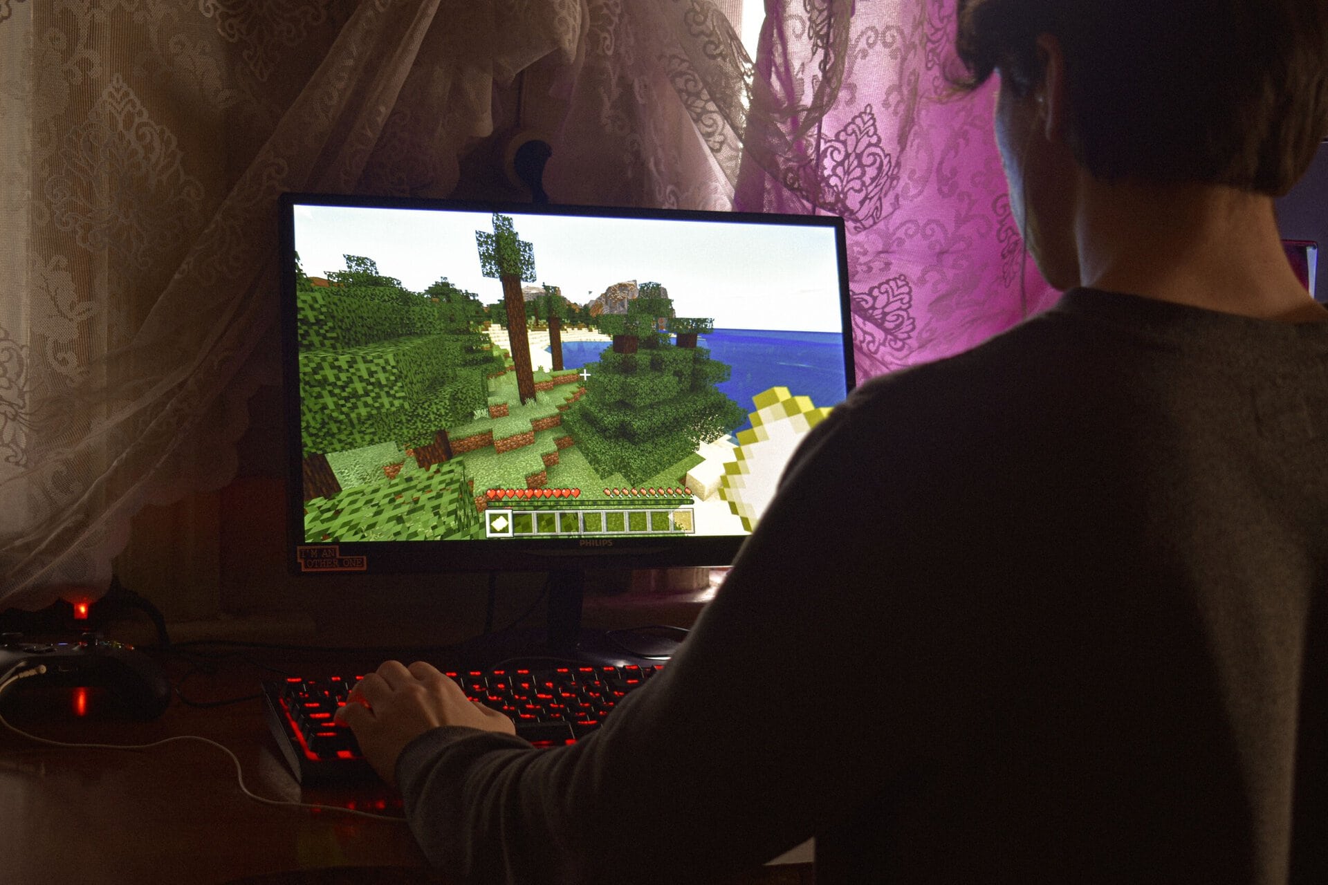 a person with short brown hair plays minecraft on a desktop computer with a red backlit keyboard near a window with lace curtains.