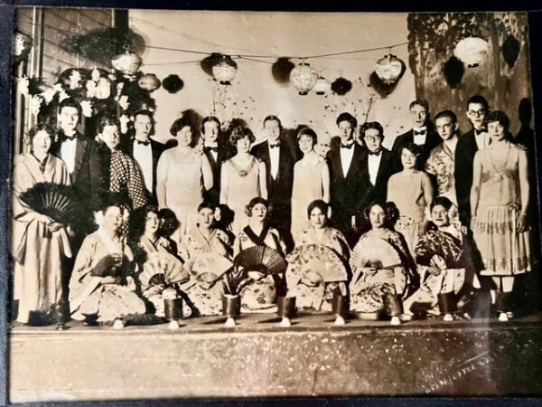 a vintage black and white group photo shows men in suits and women in dresses standing, with women in kimonos kneeling in front holding fans. lanterns hang in the background.
