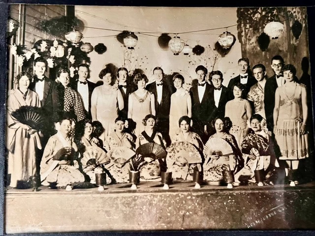 a vintage black and white group photo shows men in suits and women in dresses standing, with women in kimonos kneeling in front holding fans. lanterns hang in the background.