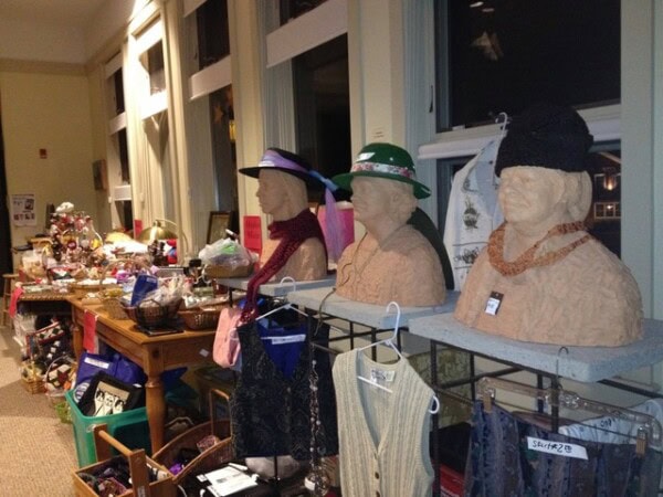 three sculpted busts wearing hats and necklaces are displayed indoors on a table next to hanging clothes, with various items and crafts arranged on tables in the background.