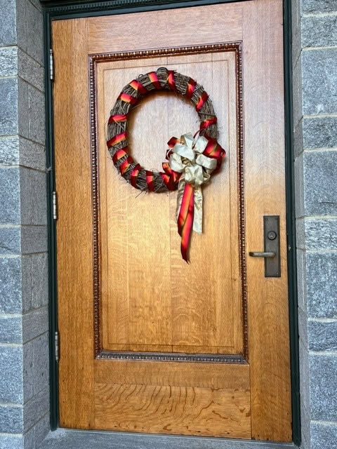 a wooden door with a brass handle features a wreath decorated with red, black, and gold ribbon and a large bow, set in a gray stone wall.