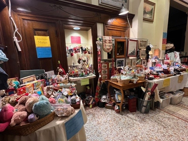 indoor market stall with tables and baskets displaying assorted toys, decorations, and gifts, set against wooden paneled walls and signs reading “meclan’s market” and “cashier.”.