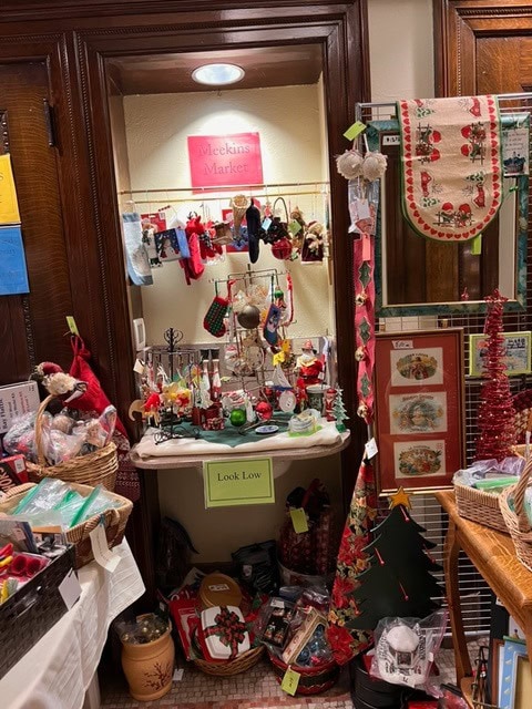 a small holiday market stall displays assorted christmas decorations, ornaments, and gifts on shelves and tables, with a "victorian market" sign above and a "look low" sign below.