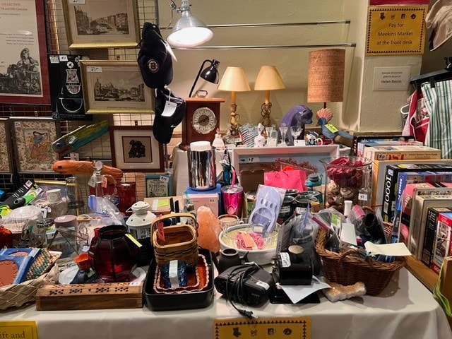 table filled with assorted secondhand items including baskets, clocks, lamps, books, framed art, mugs, kitchenware, and various knickknacks in a thrift store or flea market setting.