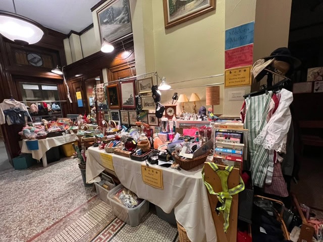 tables covered with assorted vintage items, books, clothing, and household goods are displayed for sale in a well lit indoor market area with tiled floors and wood paneled walls.