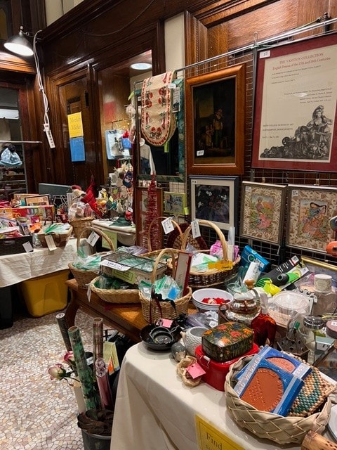 a table and shelves display assorted vintage items, crafts, and decorations in a shop with framed art and signs on the wooden paneled walls.