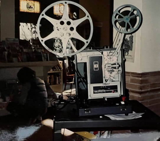 a vintage reel to reel film projector is set up on a cart in a dimly lit room with books and a person crouching nearby.
