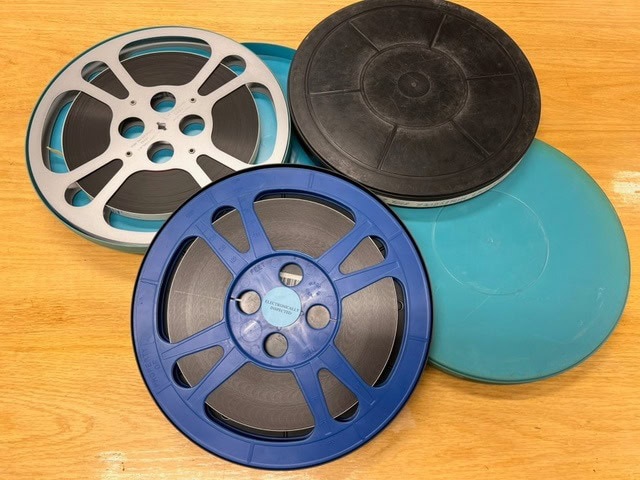 three film reels, one black, one blue, and one metal, are displayed on a light wooden surface with their matching plastic cases partially visible.