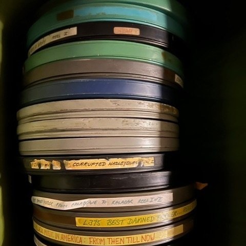 a stack of film reels with various colored lids and handwritten labels, some partially legible, arranged vertically inside a storage container.