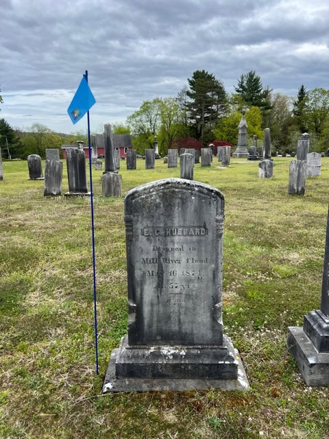 a gravestone marked "e.c. hubbard" stands in a cemetery with other headstones, a blue flag on a pole is placed next to it, trees and cloudy sky in the background.
