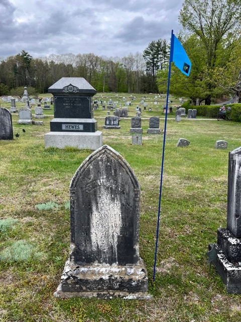 an old, weathered gravestone with faded text stands next to a blue flag in a cemetery with many other headstones under a cloudy sky.