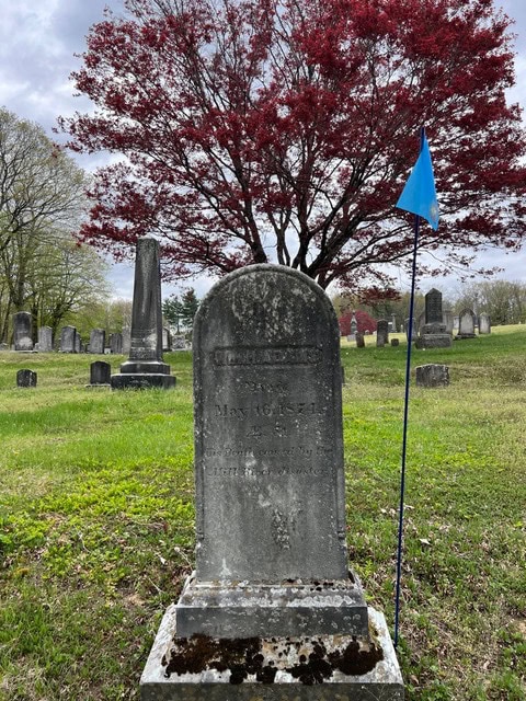 an old gravestone with weathered inscriptions stands in a cemetery, marked by a blue flag, with a red leaved tree and other headstones in the background.