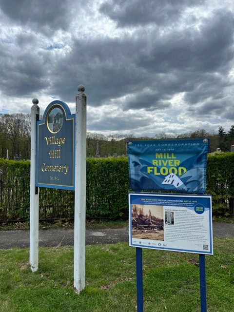 village hill cemetery sign next to informational banners about the mill river flood, with a cloudy sky and headstones visible in the background.