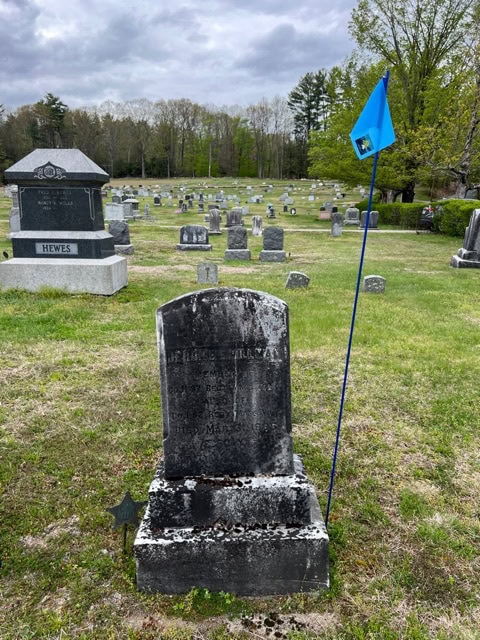 a weathered gravestone in a cemetery stands next to a blue flag on a pole, surrounded by grass and other headstones under a cloudy sky.