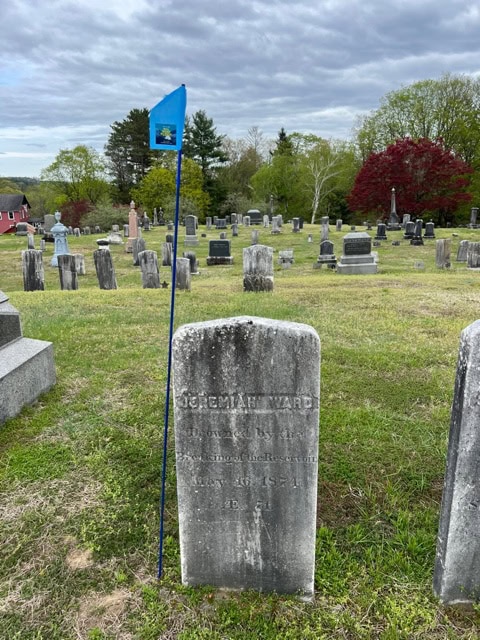 a blue flag marks the grave of jeremiah ward in a cemetery with numerous headstones and trees in the background under a cloudy sky.