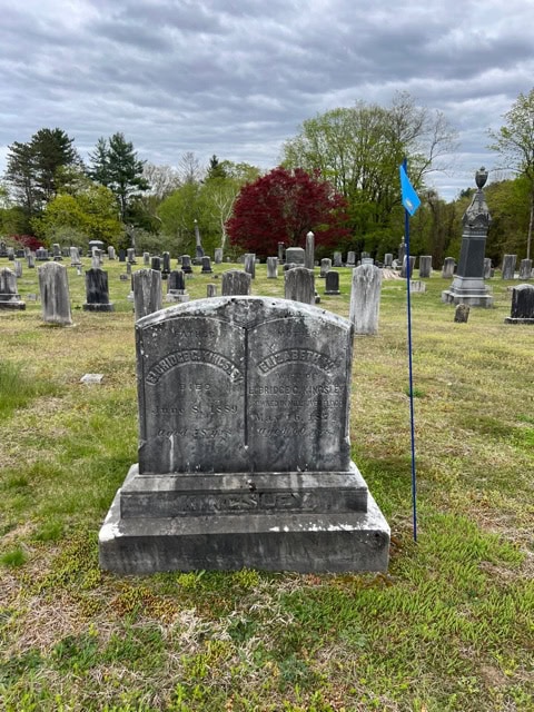 twin headstones in an old cemetery with worn inscriptions, accompanied by a blue flag on a pole, surrounded by other gravestones and trees under a cloudy sky.