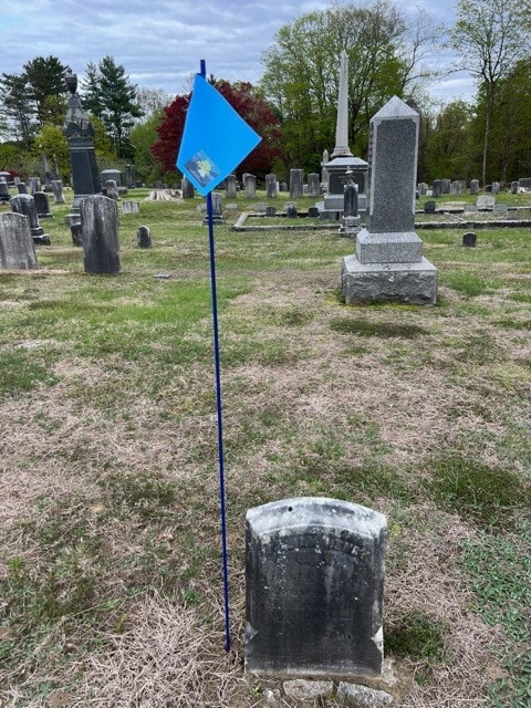 a weathered gravestone in a cemetery stands next to a blue flag on a pole; other gravestones and trees are visible in the background.