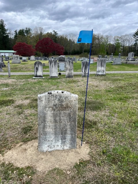 old gravestone in a cemetery, marked by a tall pole with a small blue flag attached. other headstones and trees are visible in the background under a cloudy sky.