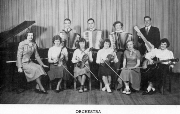 black and white photo of a school orchestra with ten members posing with instruments, including accordions, violins, piano, guitar, and upright bass, in front of a curtain.