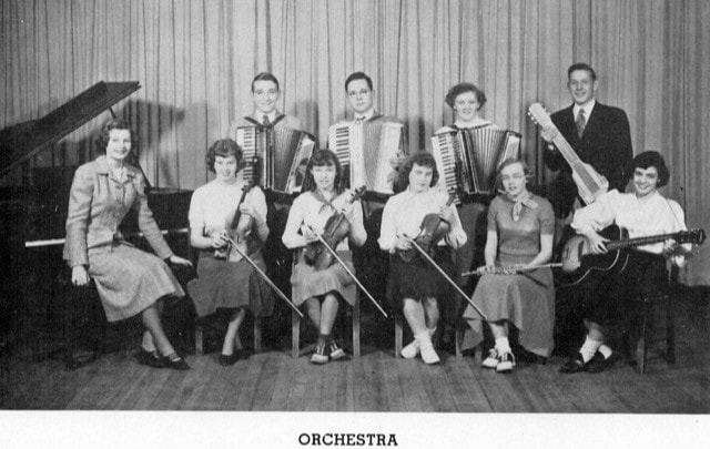 black and white photo of a school orchestra with ten members posing with instruments, including accordions, violins, piano, guitar, and upright bass, in front of a curtain.