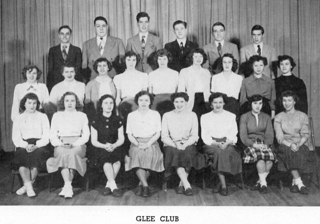 black and white photo of 24 young men and women in formal attire, posed in three rows on stage with a curtain backdrop labeled “glee club.”.