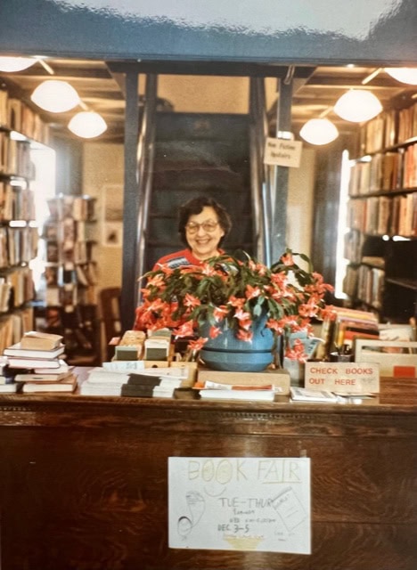 a woman stands behind a wooden library desk with books and a potted plant, surrounded by bookshelves and a staircase in the background.