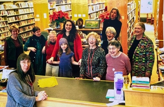 a group of women of various ages stands and smiles together in a library, with bookshelves and a checkout counter in the background.