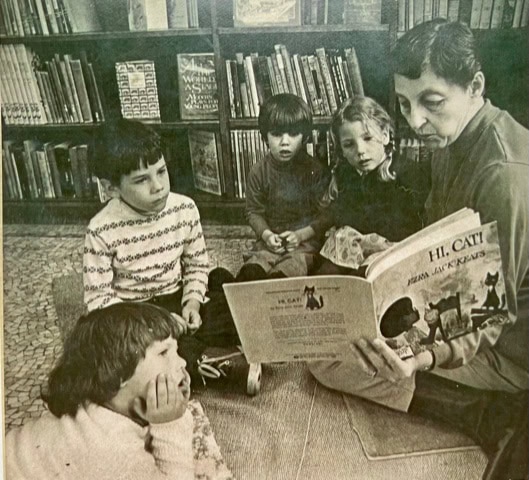 an adult reads the book "hi, cat!" to four children sitting on the floor in front of a bookshelf filled with books.