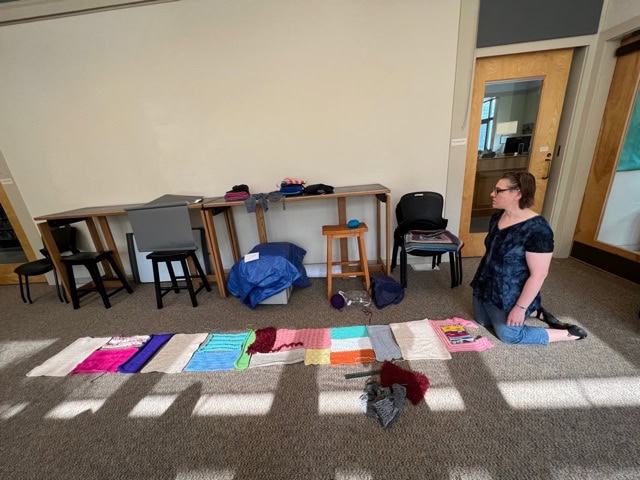 a woman kneels on the floor in a room, looking at an array of colorful knitted or crocheted items laid out on a blanket. desks, chairs, and bags are in the background.