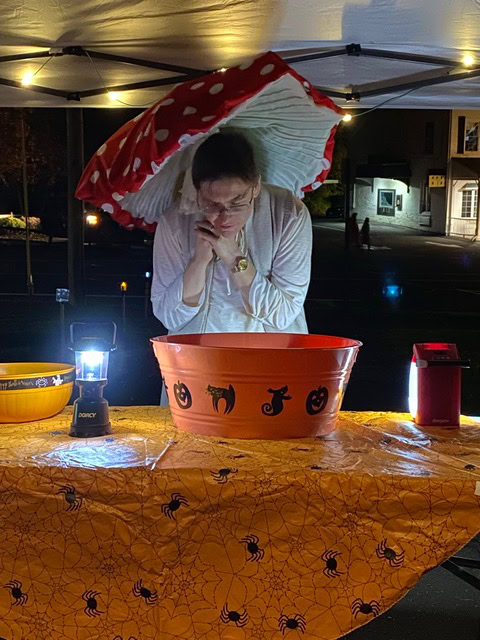 a person in a mushroom hat leans over a large orange halloween themed bowl on a table covered with a spiderweb cloth, under a canopy at night.