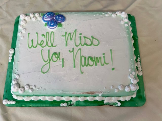 rectangular sheet cake with white icing, green border, blue and purple rose decorations, and the message "we'll miss you, naomi!" written in green frosting.