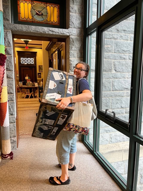 a woman wearing glasses carries a large decorated box and a tote bag while walking in a hallway with stone walls and large windows.