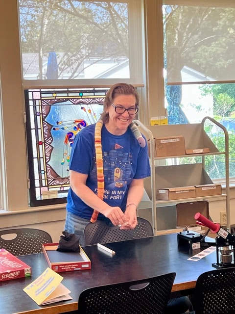 a person with glasses, wearing a snake plush around their neck, stands smiling at a table with board games and library supplies in a brightly lit room.