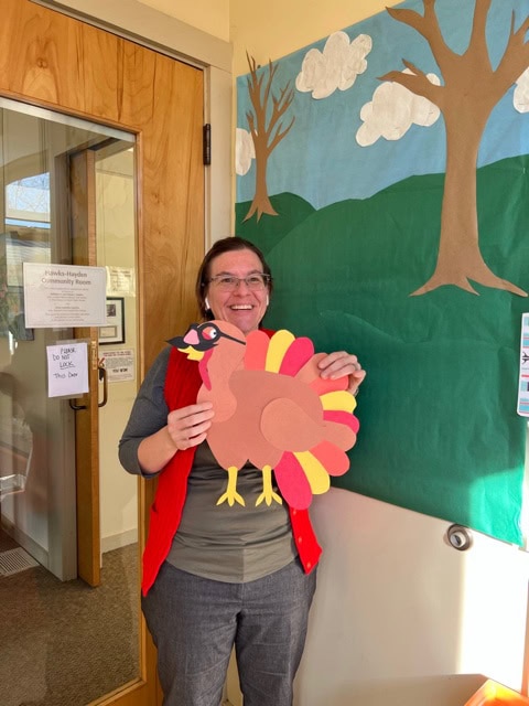a woman standing indoors holds a large, colorful paper turkey decoration and smiles at the camera, with a bulletin board featuring a tree and clouds in the background.