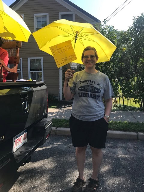 person standing outdoors holding a yellow umbrella and a sign that reads "meekins library," wearing a gray "property of library" t shirt and black shorts, next to a black vehicle.
