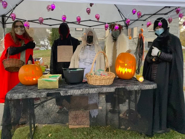 five people in halloween costumes stand behind a decorated table with carved pumpkins, baskets, and treats under a canopy with purple skull lights.
