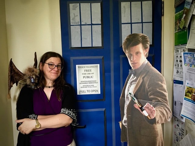 a woman stands next to a tardis door with a cardboard cutout of the eleventh doctor from doctor who; she has a bat puppet on her shoulder.