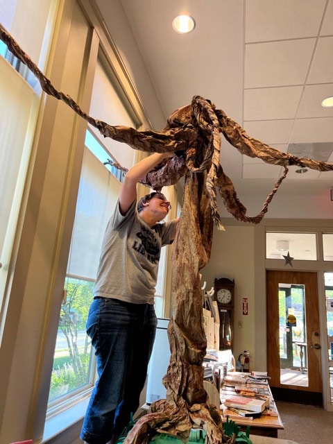 a person standing on a chair attaches long brown paper strips to a tall structure indoors, near large windows and a table with books.