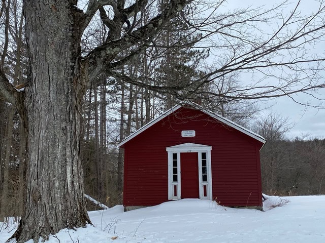 a small red building with double white doors stands in snow next to a large leafless tree; bare trees are visible in the background under a cloudy sky.