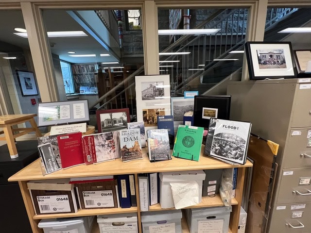 a display of books, documents, and photographs organized on shelves and a filing cabinet in an office or archive setting, with storage boxes and framed images.