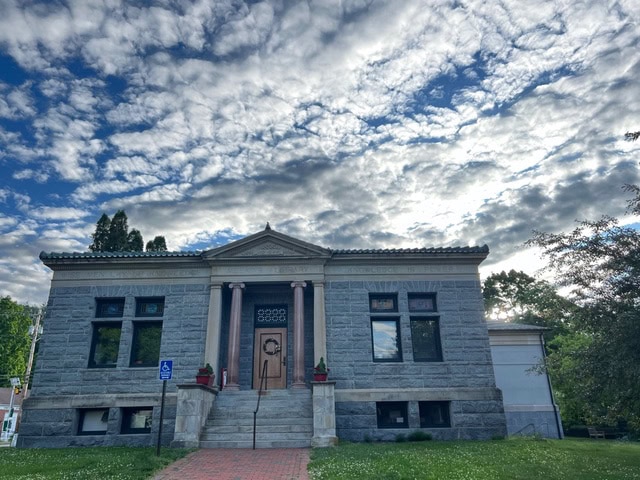 a stone building with four columns at its entrance sits under a partly cloudy sky, with a brick path leading to the front steps and greenery surrounding the structure.
