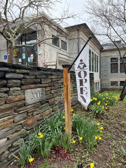 a building with a stone wall marked "2005," daffodils in bloom, and a white flag reading "open" on a flagpole near the entrance.