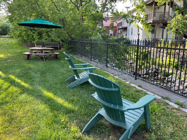 two green adirondack chairs face a black iron fence, with a wooden picnic table and green umbrella nearby on a grassy area next to trees and houses.