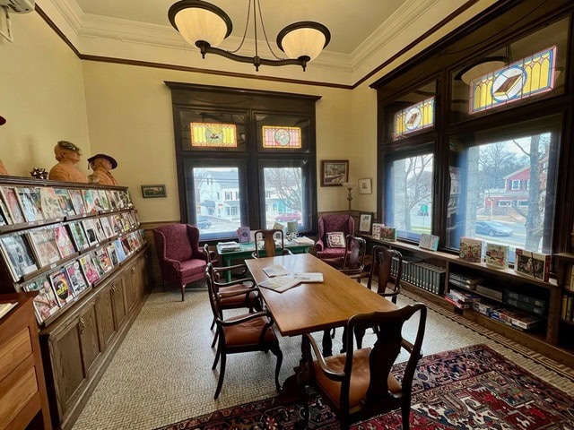 a library reading room with a wooden table and chairs, magazines on display, bookshelves, large windows with stained glass, and natural light filling the space.