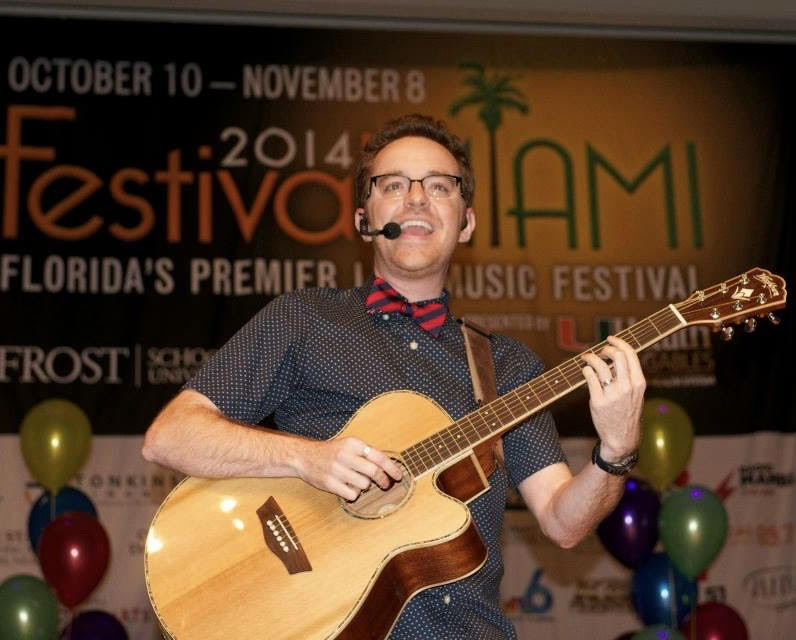 a man wearing glasses, a headset mic, and bow tie plays an acoustic guitar on stage at a music festival with colorful balloons in the background.