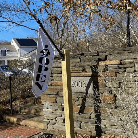 a white "open" flag hangs from a post near a stone wall engraved with the number 2003, with a house and trees in the background.