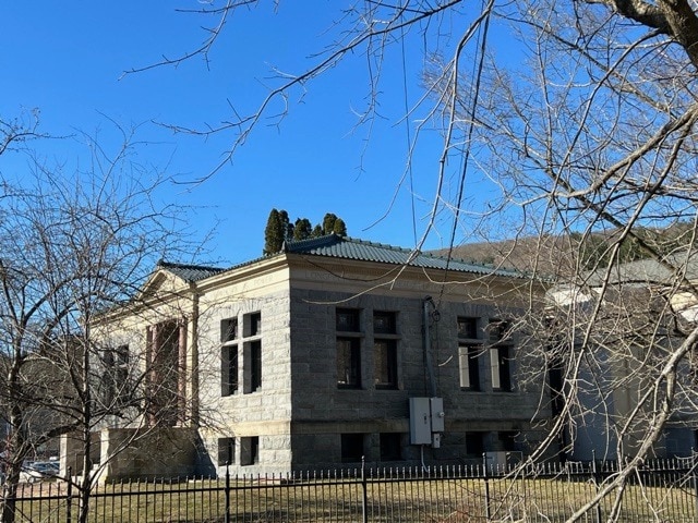 a stone building with large windows and a green roof, surrounded by leafless trees and a black metal fence under a clear blue sky.