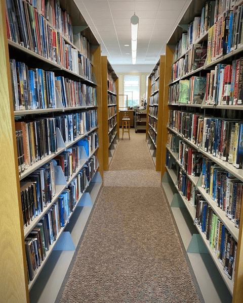 library aisle lined with bookshelves on both sides, leading to a window with a wooden stool at the end of the walkway.