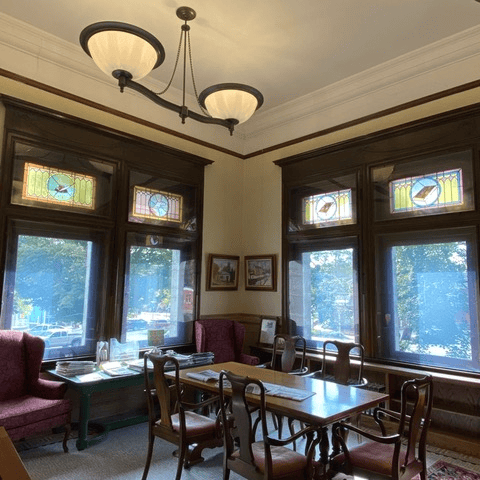 a sunlit room with stained glass windows, wooden trim, a chandelier, upholstered chairs, and a dining table with newspapers and magazines.