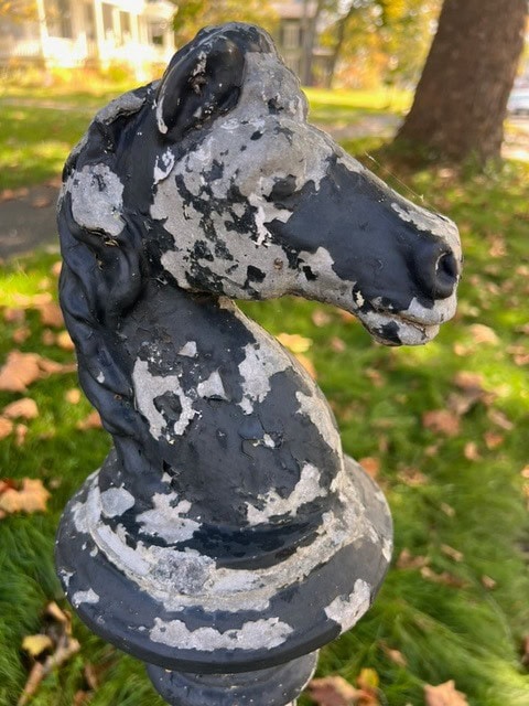 a weathered black horse head hitching post with peeling paint stands outdoors on grass with fallen leaves in the background.